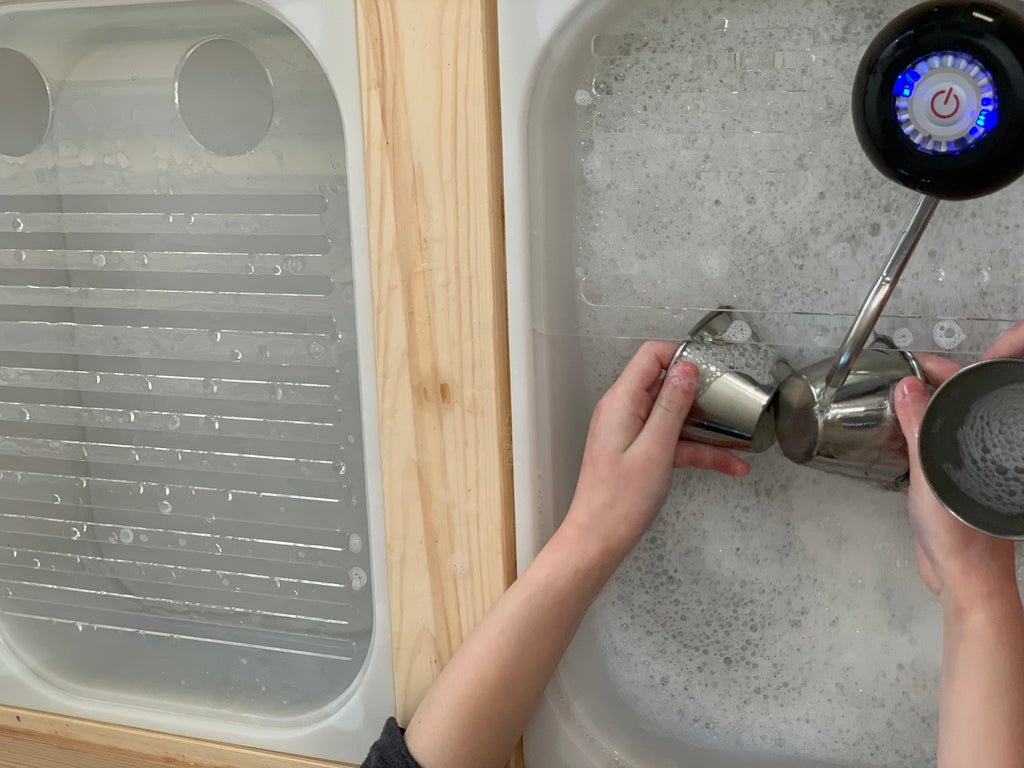 Child's hands washing small metal cups in wooden play sink insert with dish rack beside, filled with bubbly water