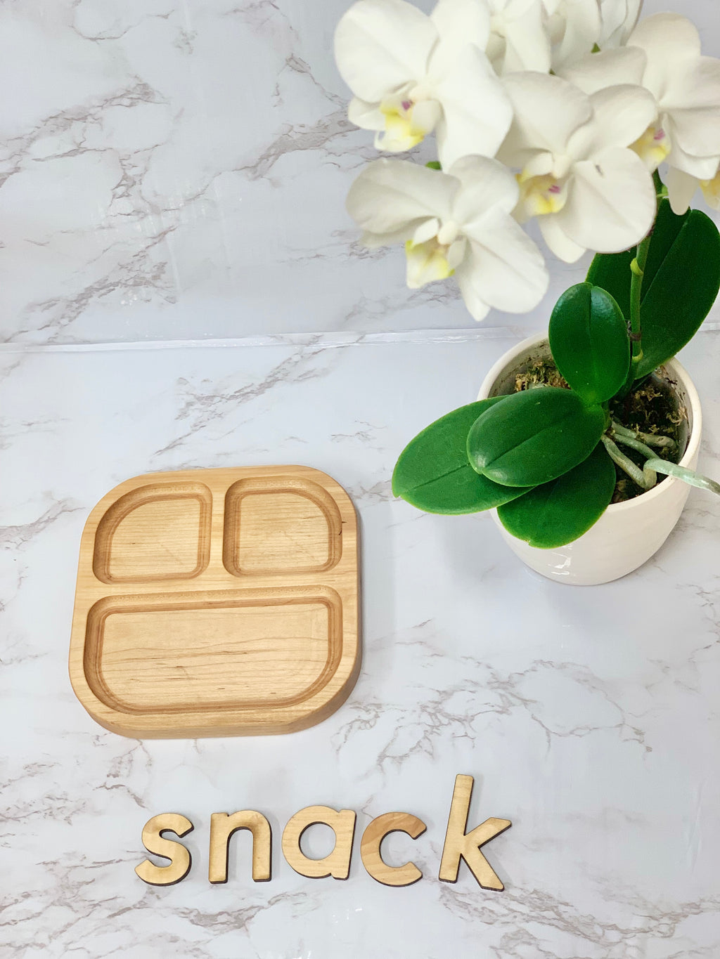Square wooden sensory tray with three compartments displayed on marble surface near white orchid plant and wooden letters spelling snack