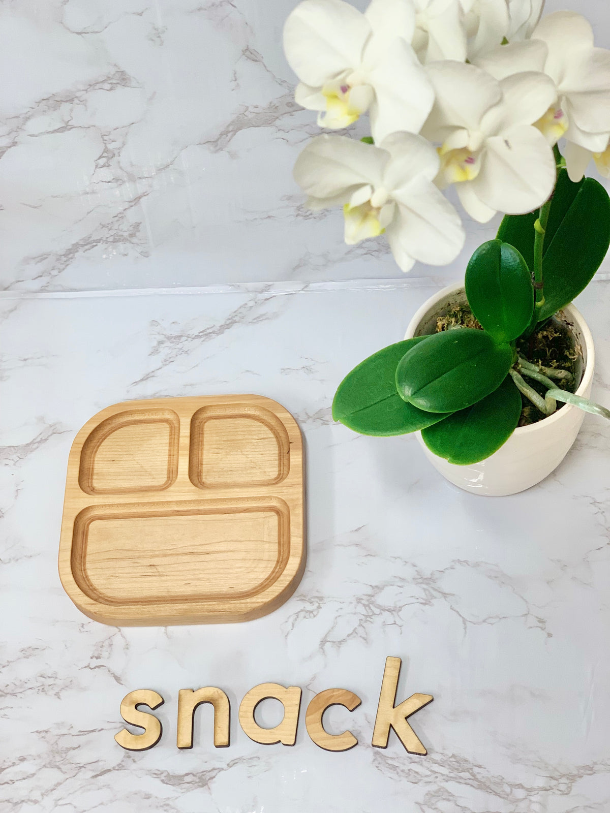 Square wooden sensory tray with three compartments displayed on marble surface near white orchid plant and wooden letters spelling snack