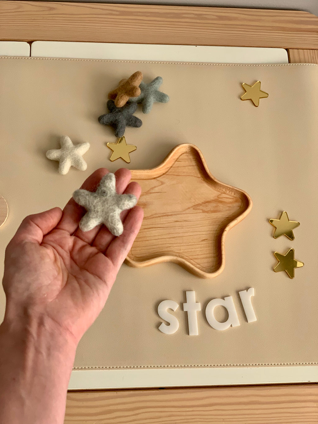 Hand holding neutral gray felt star near wooden star-shaped tray with white letters and gold star decorations on beige surface