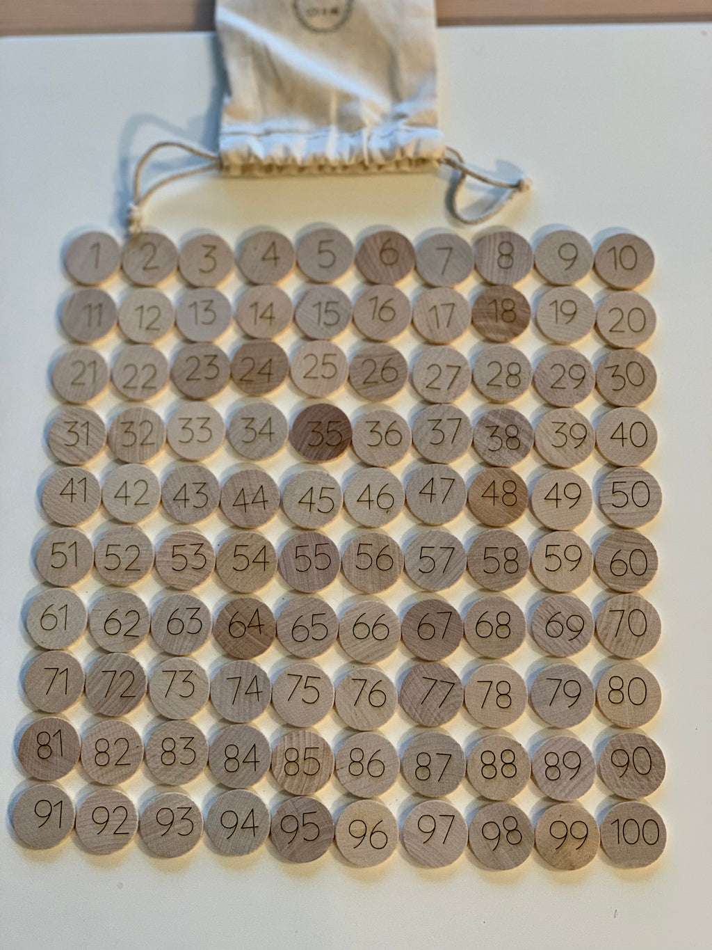 Set of 100 laser engraved wooden number discs from 1 to 100 arranged in rows with drawstring cloth bag on white surface