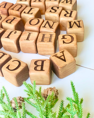 Natural 1.5-inch wooden alphabet blocks with engraved capital letters scattered near green pine branches on white surface
