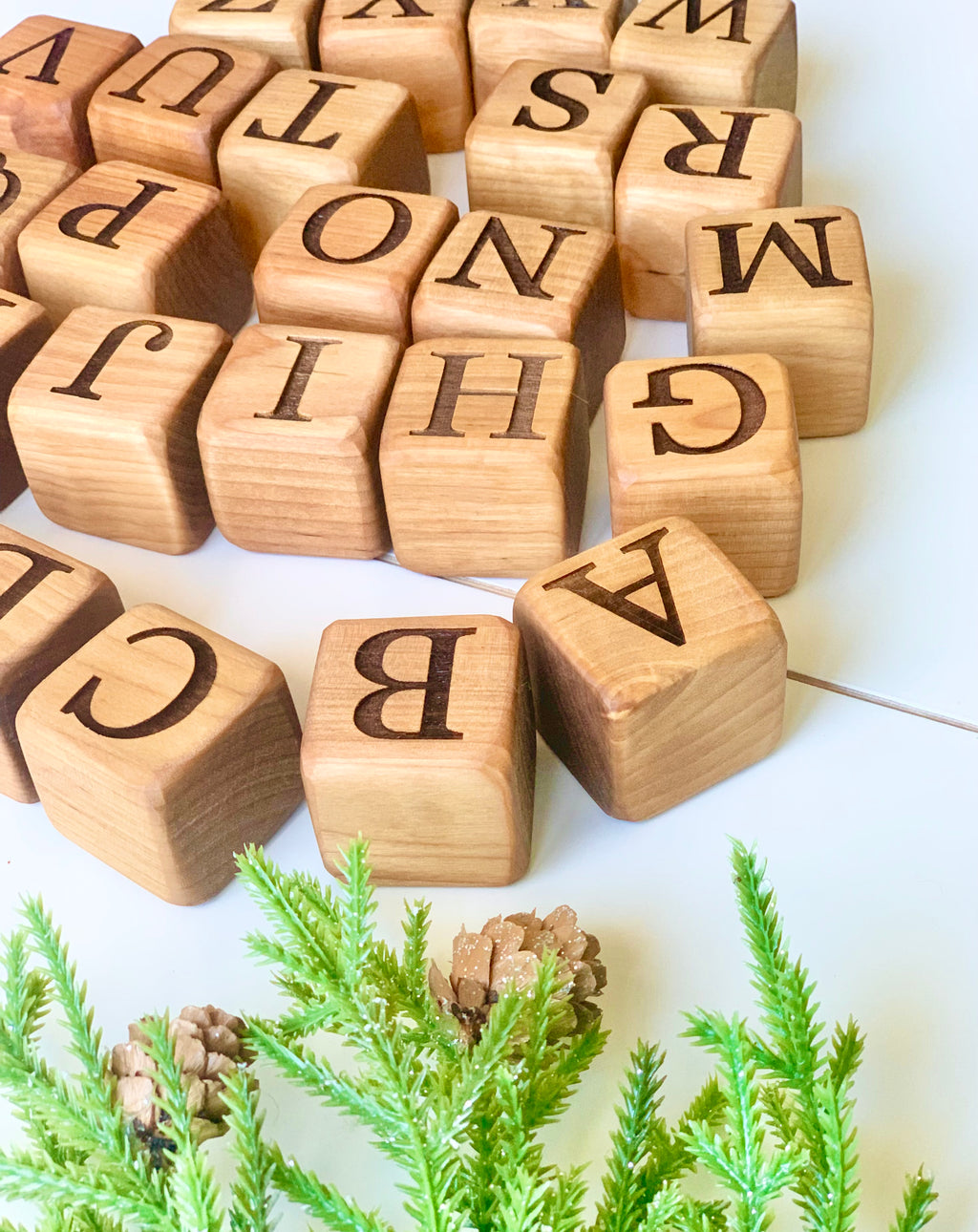 Natural 1.5-inch wooden alphabet blocks with engraved capital letters scattered near green pine branches on white surface
