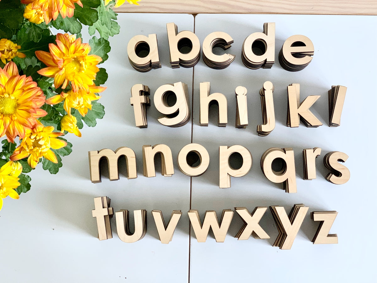 Natural wooden lowercase alphabet letters arranged in rows on white surface near yellow flowers