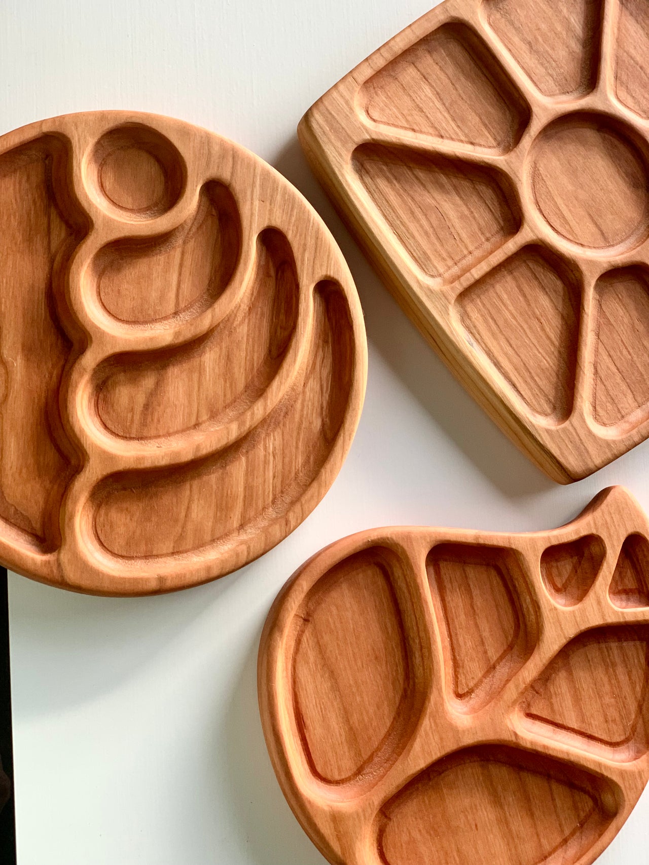 Three wooden sensory trays in round, leaf, and flower shapes with multiple carved compartments on white surface