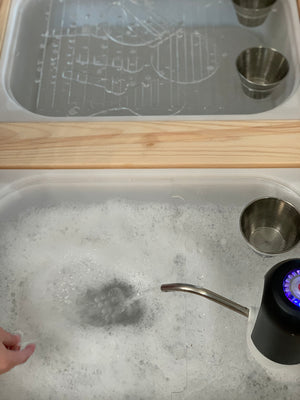 Play kitchen sink insert filled with soapy water and small stainless steel cups on wooden surface