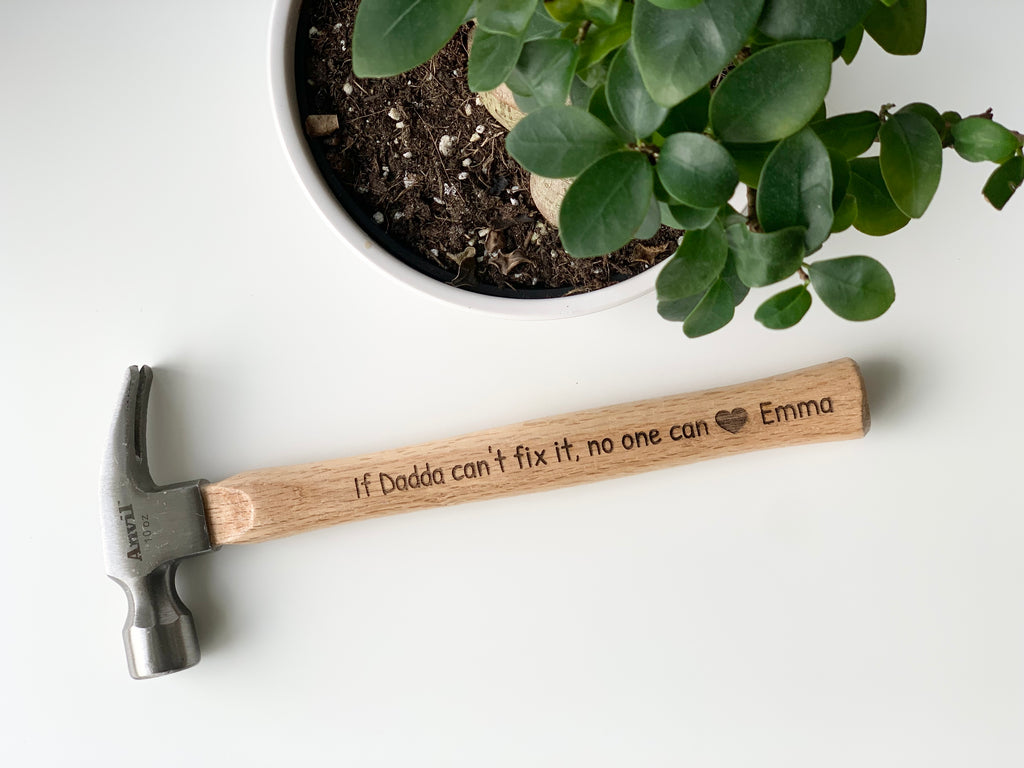 Natural wooden hammer with custom laser engraved message and heart symbol near green potted plant on white surface