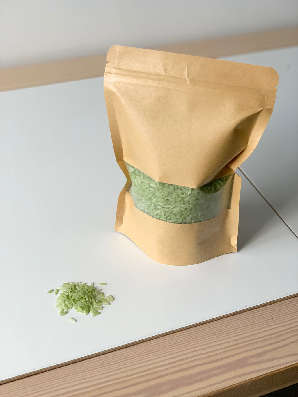 Brown kraft resealable pouch filled with light green sensory rice spilled on white and wood surface