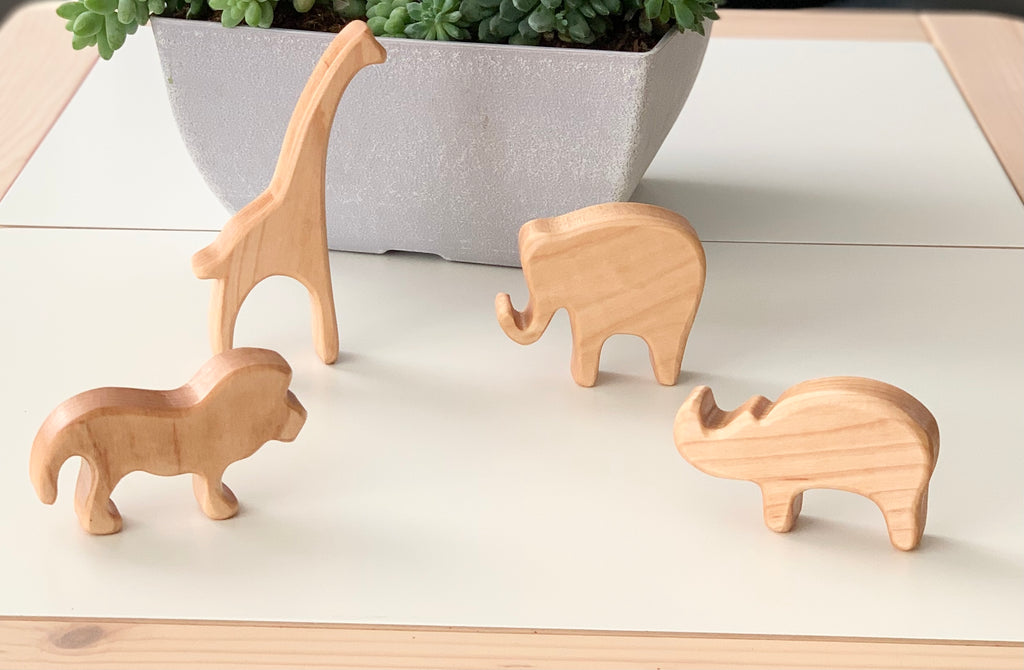 Natural wooden safari animal toy figurines including giraffe, elephant, lion, and rhinoceros on white surface with planter background