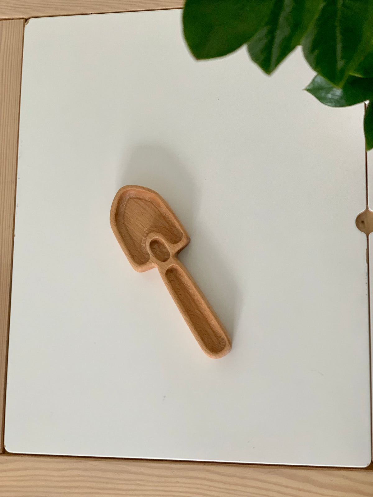 Small wooden shovel-shaped sensory tray on white surface with green plant leaves in corner