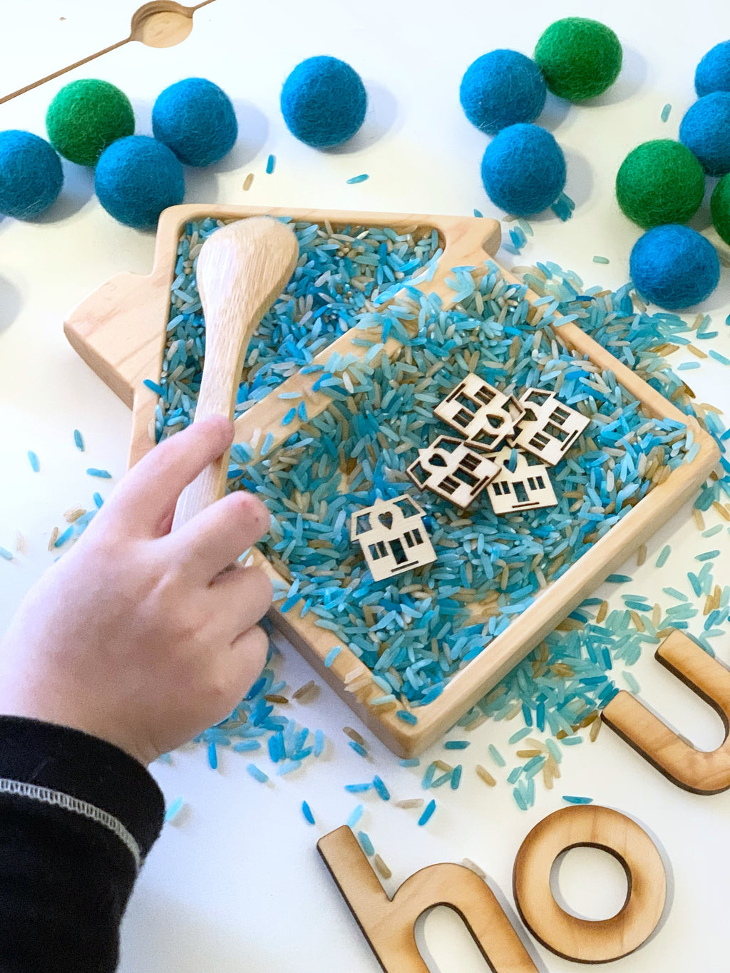 Natural wooden house-shaped sensory tray filled with blue rice and small wooden house pieces with child's hand holding wooden spoon