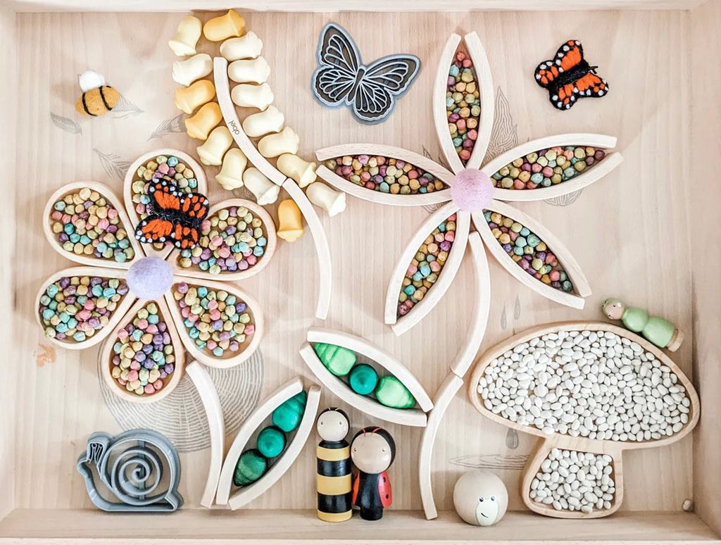 Wooden sensory tray with rain drop plates filled with colorful beans, wooden mushroom, butterflies, bees, and snail figures on wood background