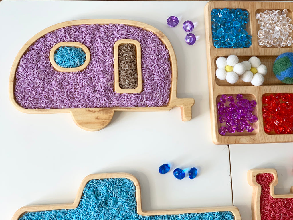 Camper-shaped wooden sensory tray filled with colored rice next to wooden tray with colorful sensory play stones and balls