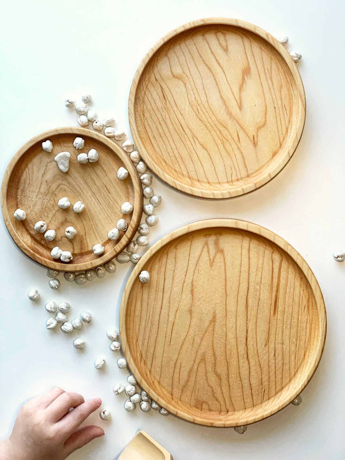 Three natural wooden round nesting plates arranged on white surface with scattered small white textured balls and child's hand