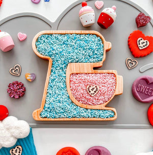 Wooden mixer-shaped sensory tray filled with blue and pink rice surrounded by colorful Valentine's Day-themed decorations
