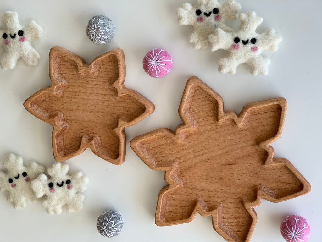 Two natural wooden snowflake-shaped small plates surrounded by soft snowflake toys and decorative balls on white surface